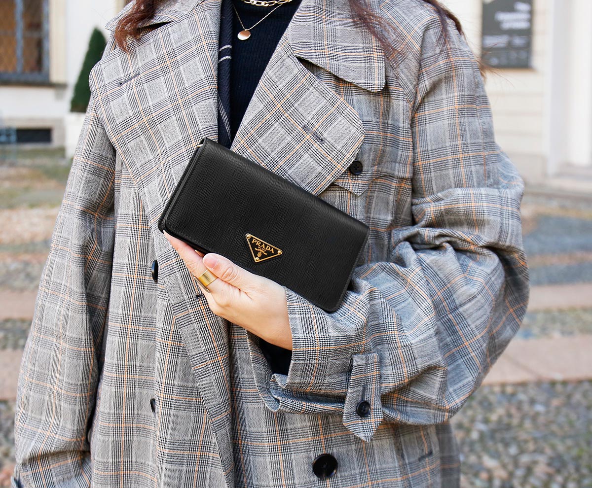 Person wearing a plaid coat holding a black Prada wallet outdoors.